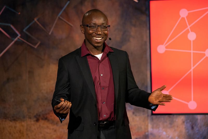 A male presenter smiling at the audience with hands in an outstretched open gesture