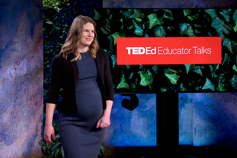 A pregnant presenter looking toward the audience, beside a screen displaying "TED-Ed Educator Talks"