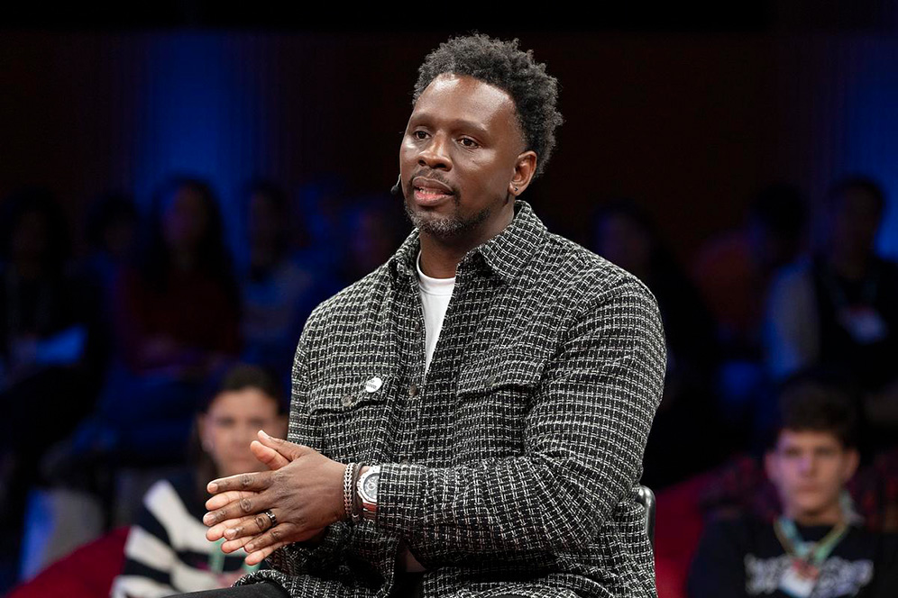 A man seated onstage, wearing a black-and-white patterned jacket, speaks with his hands clasped while an audience watches